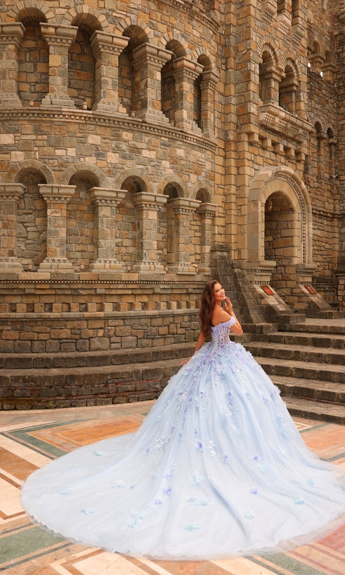 A woman wears the Amarra 54231 3D Floral Quinceañera Dress in light blue, standing on a stone courtyard before a grand, ornate stone building.